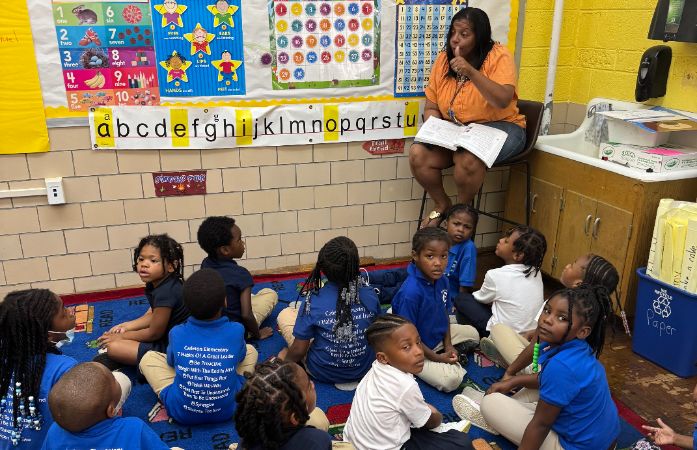  Students sitting on rug listening to teacher reading a story.